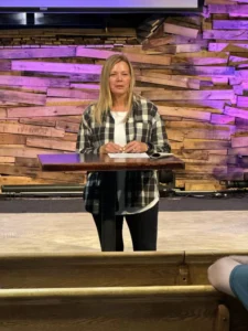 Rachel speaking behind a podium at a church with a wooden backdrop lit by purple lights.